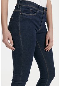 Jeans de corte slim en azul oscuro, hechos de denim con costuras en color cobre. Cuenta con cinco bolsillos y un diseño de cintura alta. Textura suave.