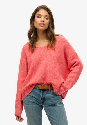 Roze, oversized trui met een V-hals en geribbelde manchetten, gemaakt van zacht textuurmateriaal. Gecombineerd met lichtblauwe jeans en een bruine riem.