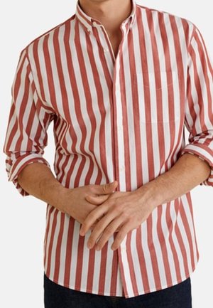 Mężczyzna w koszuli z długim rękawem z czerwono-białymi pionowymi paskami, rękawy podwinięte, ręce splecione z przodu.