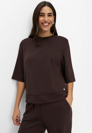 Lachende vrouw met donker haar draagt een donkerbruine korte mouw top en bijpassende broek, staand tegen een eenvoudige witte achtergrond.