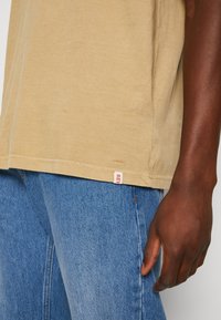 Beige cotton t-shirt with short sleeves and a straight hem, featuring a small red logo tag at the bottom and worn with blue denim jeans.