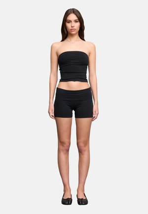 Jeune femme debout face à l’objectif portant un haut noir sans bretelles, un short noir ajusté et des chaussures noires à orteils fendus sur un fond uni.