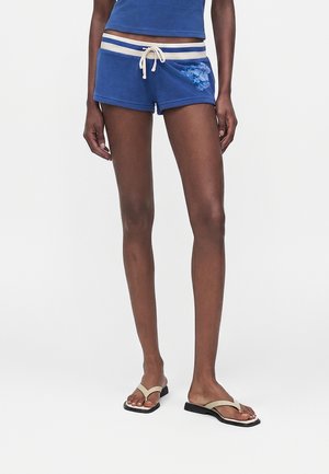 Femme portant un short bleu à cordon avec imprimé floral et des sandales plates beiges, debout sur un fond blanc.