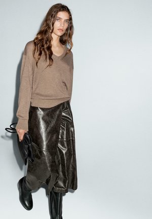 Maglione marrone lavorato a maglia con scollo a V, abbinato a una gonna in pelle nera scura e stivali neri fino al ginocchio. La modella tiene una borsa nera.