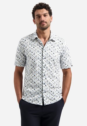 Mann mit lockigem Haar, der ein helles kurzärmeliges, gemustertes Hemd mit Knöpfen und dunkle Hosen trägt, Hände in den Taschen, neutraler Hintergrund.