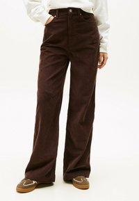 Pantaloni a vita alta in corduroy marrone con gamba larga, strisce verticali testurizzate e tasche anteriori, abbinati a sneakers marroni con dettagli bianchi.