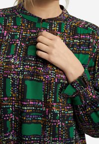 Camicia con collo e bottoni in un vivace motivo a pois neri, verdi e rosa con tessuto strutturato; presenta maniche arricciate con polsini arruffati.