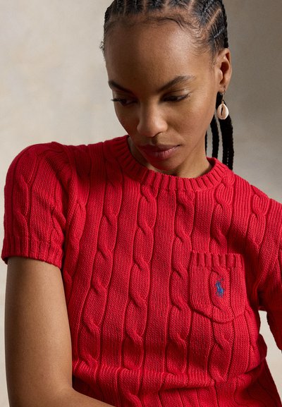 Kobieta z warkoczami typu cornrow, ubrana w czerwony, krótki sweter z grubym splotem z małą kieszonką i kolczykiem w kształcie muszli, patrząca w dół.