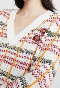 Jersey de punto en blanco con rayas rojas, verdes y naranjas en forma de V. Cuenta con botones decorativos con forma de flores. Material texturizado.