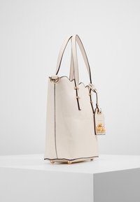 Beige scalloped tote bag made of textured material, featuring gold hardware, a decorative tag, and four round feet for stability.