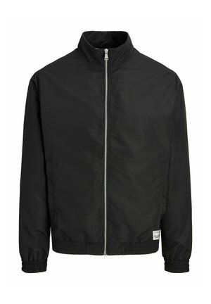 Veste noire à fermeture éclair avec col haut, poignets et ourlet élastiques, et une petite étiquette blanche rectangulaire près de l'ourlet inférieur à l'avant côté droit.