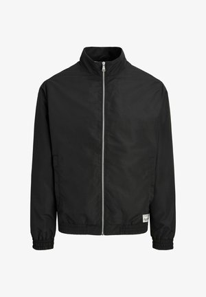 Veste noire à fermeture éclair avec col haut, poignets et ourlet élastiques, et une petite étiquette blanche rectangulaire près de l'ourlet inférieur à l'avant côté droit.