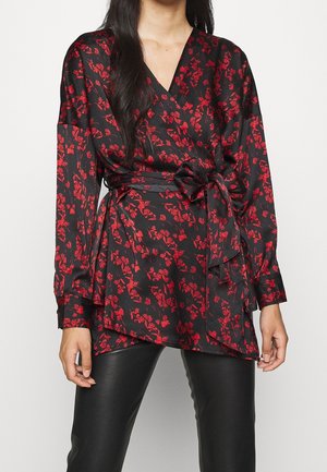 Kobieta w czarnej bluzce z nadrukiem czerwonych kwiatów, owiniętej i zawiązywanej z boku, w połączeniu z czarnymi skórzanymi spodniami.