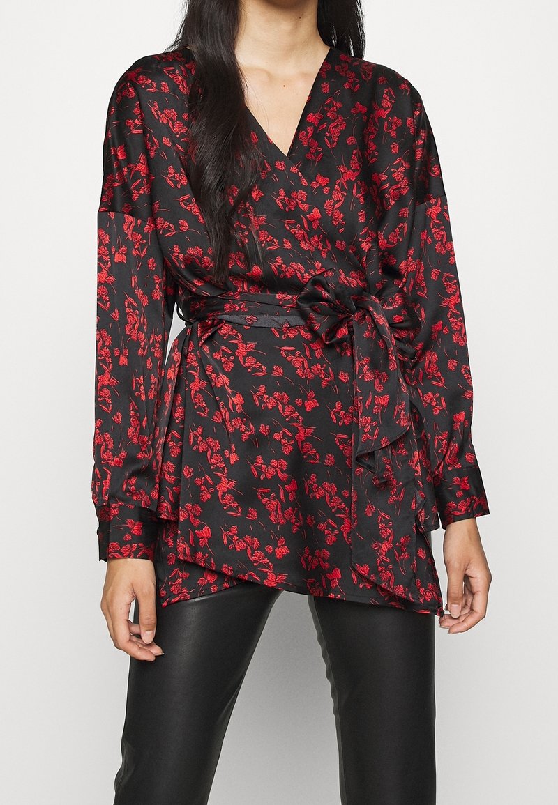 Femme portant un chemisier noir enveloppant avec un imprimé floral rouge et un lien latéral, associé à un pantalon en cuir noir.
