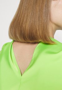 Woman with straight, shoulder-length light brown hair wearing a bright green satin top with a V-shaped cutout at the neckline back.