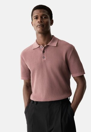 Polo de manga corta acanalado en rosa empolvado con cuello y placket de tres botones. Usado con pantalones oscuros, manos en los bolsillos.