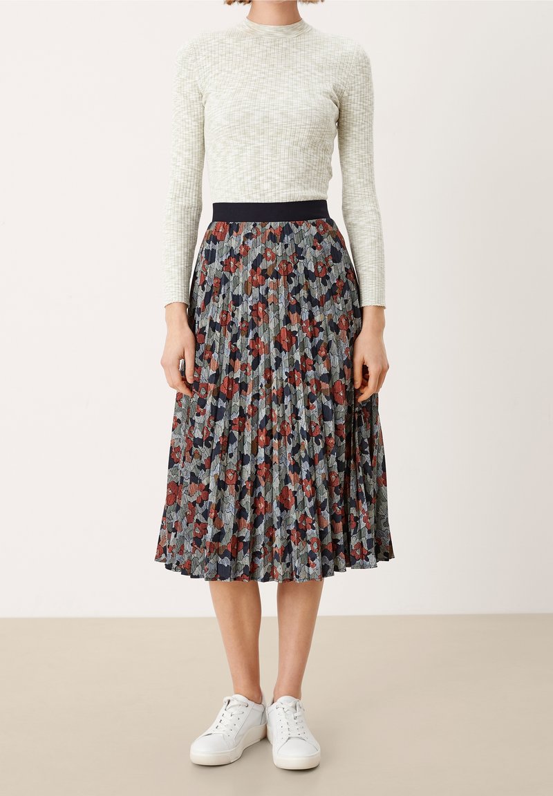 Haut blanc à manches longues avec un motif texturé, associé à une jupe midi plissée ornée d'un motif floral en rouge, marine et vert. Baskets blanches.