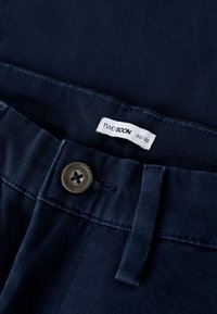 Granatowe spodnie o gładkiej teksturze, z okrągłym, ciemnym guzikiem i metką z napisem "TWO SOON 32/32" przy pasku.