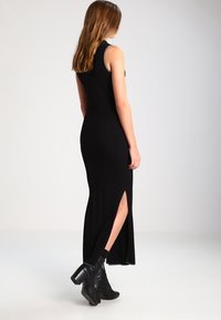 Schwarzes, geripptes Maxikleid mit seitlichem Schlitz, ärmellosem Design, tailliertem Schnitt und subtiler Glanz. Tragen mit schwarzen Ankle Boots.