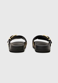 Sandali in pelle nera con suola piatta, una fascia larga e dettagli con fibbia in tono oro. La texture appare liscia e il design è minimalista.