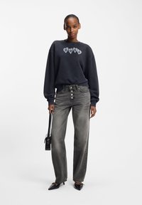 Oversized svart sweatshirt med "HUGO"-logotyp, kombinerad med gråa högmidjade jeans med knapplås och svarta spetsiga klackskor.