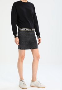 Sudadera negra con el texto "te pliego así" en la cinturilla, combinada con una falda de mezclilla gris y zapatillas blancas. Diseño de atuendo casual.