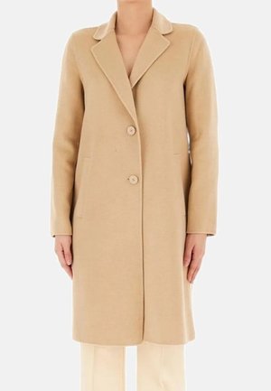 Beige knielange jas met twee knopen, reverskraag met inkeping, lange mouwen en zijzakken, gedragen over crèmekleurige broek.