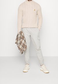 Helles beiges Strickoberteil mit einem kleinen gelben Bananenemblem, kombiniert mit hellgrauen Cargo-Hosen und cremefarbenen Sneakers.