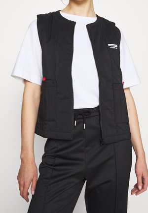 Person trägt eine schwarze Zip-Up-Weste mit Fronttaschen über einem weißen kurzärmligen Shirt und schwarzen Hosen mit Kordelzug.
