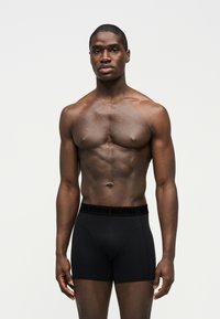 Homme musclé debout sur fond blanc, portant un boxer Björn Borg noir, regardant devant avec une expression neutre.