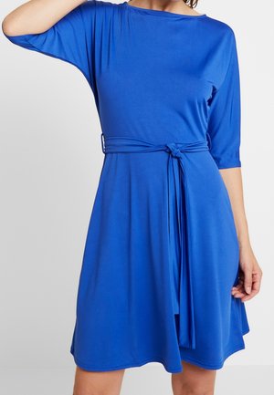 Femme portant une robe bleu vif mi-longue, manches trois-quarts, avec une ceinture nouée à la taille, debout devant un fond uni.