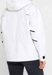 Veste imperméable blanche avec une capuche, présentant des accents noirs et des zips. Tissu texturé, poignets élastiques et design à coupe décontractée.