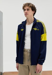 Laivastonsininen vetoketjullinen verkkaritakki keltaisin korostein ja Adidas-logo. Varustettu resorikätevine ja urheilullisella designilla. Päällä valkoinen paita.