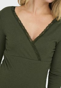 Olivgrön ribbad topp med djup V-ringning, försedda med fina mörka spetsdetaljer längs ringningen och ärmarna. Slät textur.