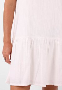 Robe en coton blanc avec un tissu léger et texturé, dotée d'un ourlet à volants superposés. La vue en gros plan montre le bras de la robe et les détails des volants.