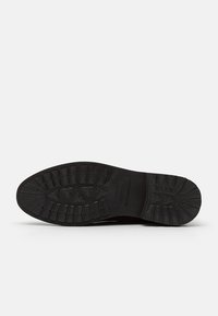 Semelle en caoutchouc noire d'une chaussure habillée avec des motifs de bande de roulement et le nom de la marque embossé au centre sur un fond blanc.