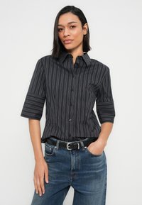 Femme portant une chemise noire à rayures fines avec manches jusqu'au coude et un jean bleu, debout avec une main dans la poche.