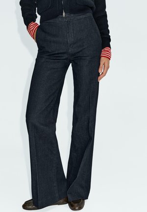 Persoon in hoge taille donkere wijde broek, zwarte trui met rode en witte gestreepte manchetten, en brown loafers, staand met één hand in de zak.