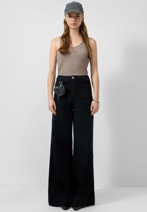 Frau trägt eine graue Mütze, ein taupefarbenes, strukturiertes Tanktop und schwarze, weit geschnittene, hoch taillierte Hosen mit einem kleinen, herzförmigen Beutel daran befestigt.