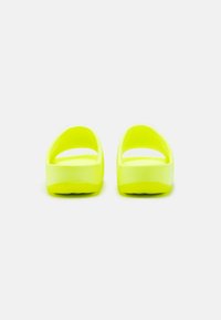Neon dzeltenas slēpņu sandales ar biezām zolēm, skatītas no aizmugures uz balta fona.