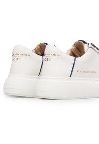 Weiße Sneakers mit einer glatten Lederoberfläche, goldener Markenkennzeichnung, einer dicken strukturierten Sohle und einem kontrastierenden blauen Akzent an der Rückseite.