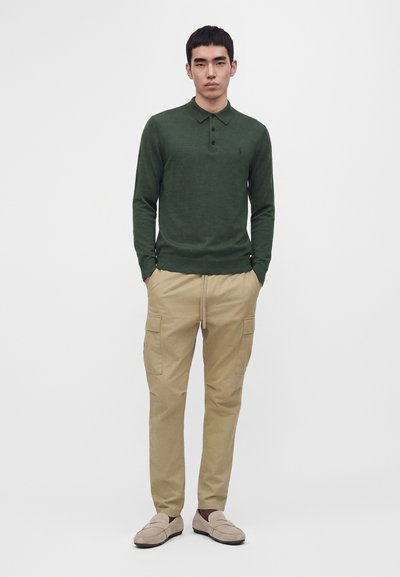 Homme portant un polo vert foncé à manches longues, un pantalon cargo beige avec des poches et des chaussures beige à enfiler, debout les mains dans les poches.