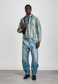 Šviesiai mėlyna džinsinė striukė ir kelnės su nublukusiomis juostelėmis ir susidėvėjimo detalėmis. Modelis laiko juodą rankinę, dėvi marškinius ir kaklaraištį.