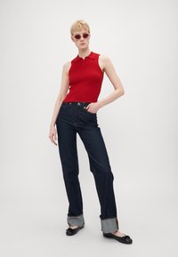 Rød ribbet ermeløs polo-topp, mørk denim høy midje bukser med brettede lyseblå mansjetter, og svarte flate sko. Modell som bærer rosa solbriller.