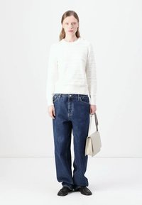 Hvit ribbet genser med rund hals, paret med løse blå denimjeans. Modellen holder en liten lysgrå veske og har på seg svarte sko.