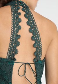 Dunkelgrünes Spitzen-Haltertop mit komplizierten Häkelbesätzen und floralen Motiven, verstellbarem Bindeschluss und strukturierten Rückendetails.