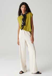 Maglietta corta verde oliva, pantaloni bianchi a gamba larga e una sciarpa a righe nere e crema con plissettature. Tessuto liscio, vestibilità rilassata e design minimalista.