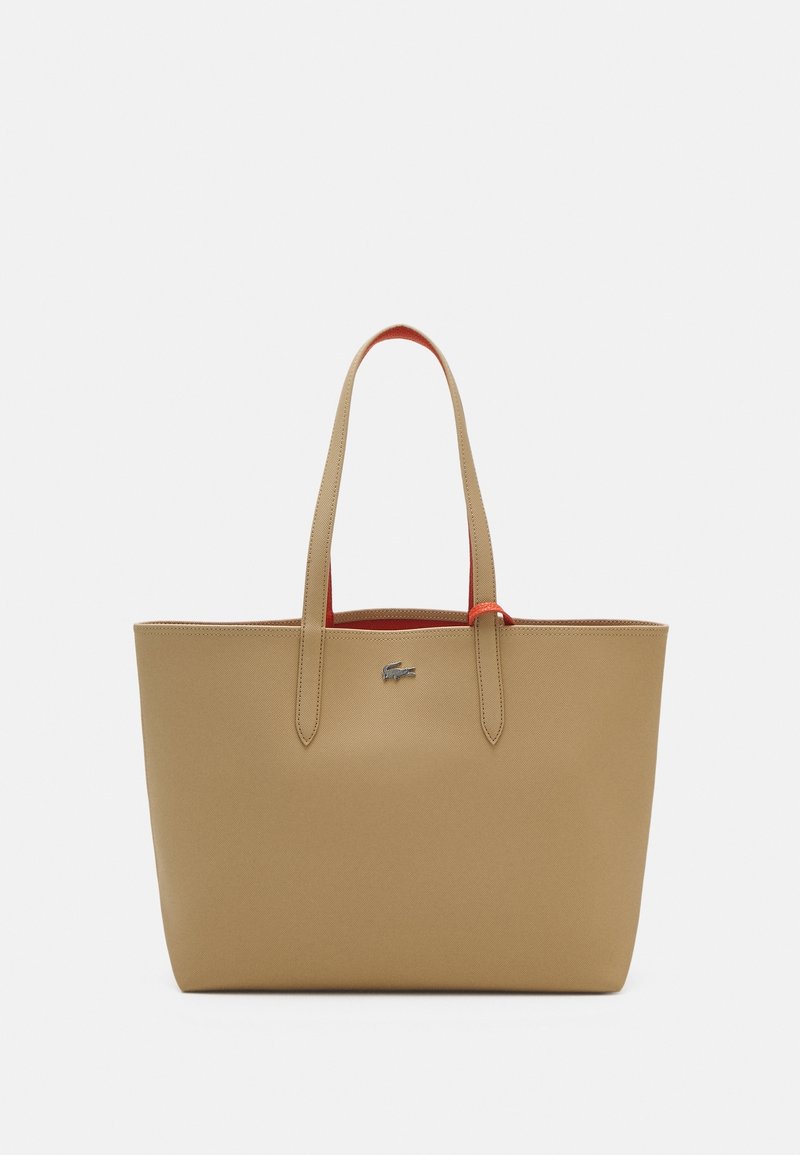 Grand sac fourre-tout couleur fauve avec longues poignées et petit logo crocodile argenté sur le devant, affichant un léger détail intérieur rouge.