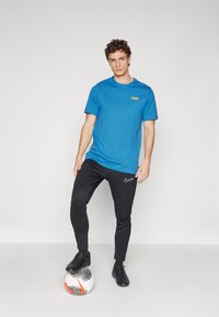 Camiseta de algodón azul con un pequeño logo en el pecho, combinada con pantalones de chándal negros que presentan el swoosh de Nike, usados con zapatillas deportivas negras.