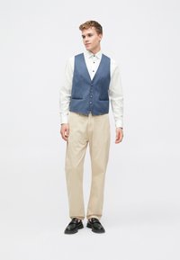 Ανδρική εμφάνιση με μπλε τζιν γιλέκο, λευκό πουκάμισο, ανοιχτό μπεζ παντελόνι και μαύρες παντόφλες, τοποθετημένη σε μια απλή φόντο.